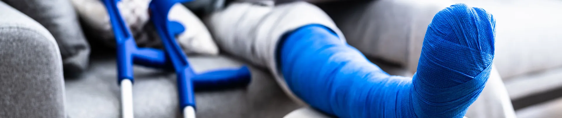 Een man met een gebroken been in blauw gips zittend op de bank met zijn been omhoog en krukken naast de bank