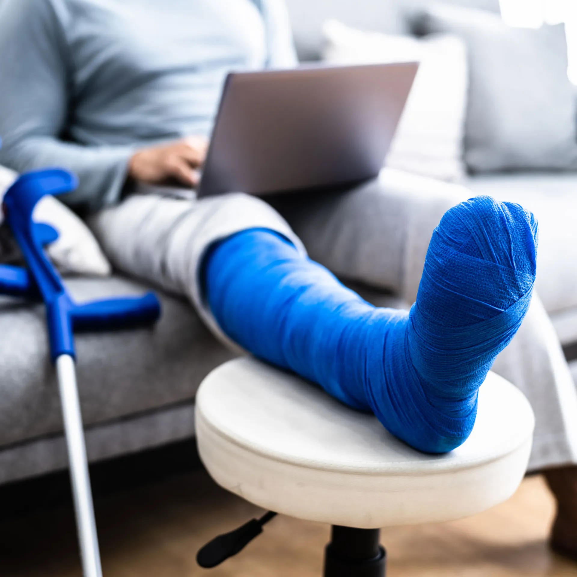 Een man met een gebroken been in blauw gips zittend op de bank met zijn been omhoog en krukken naast de bank